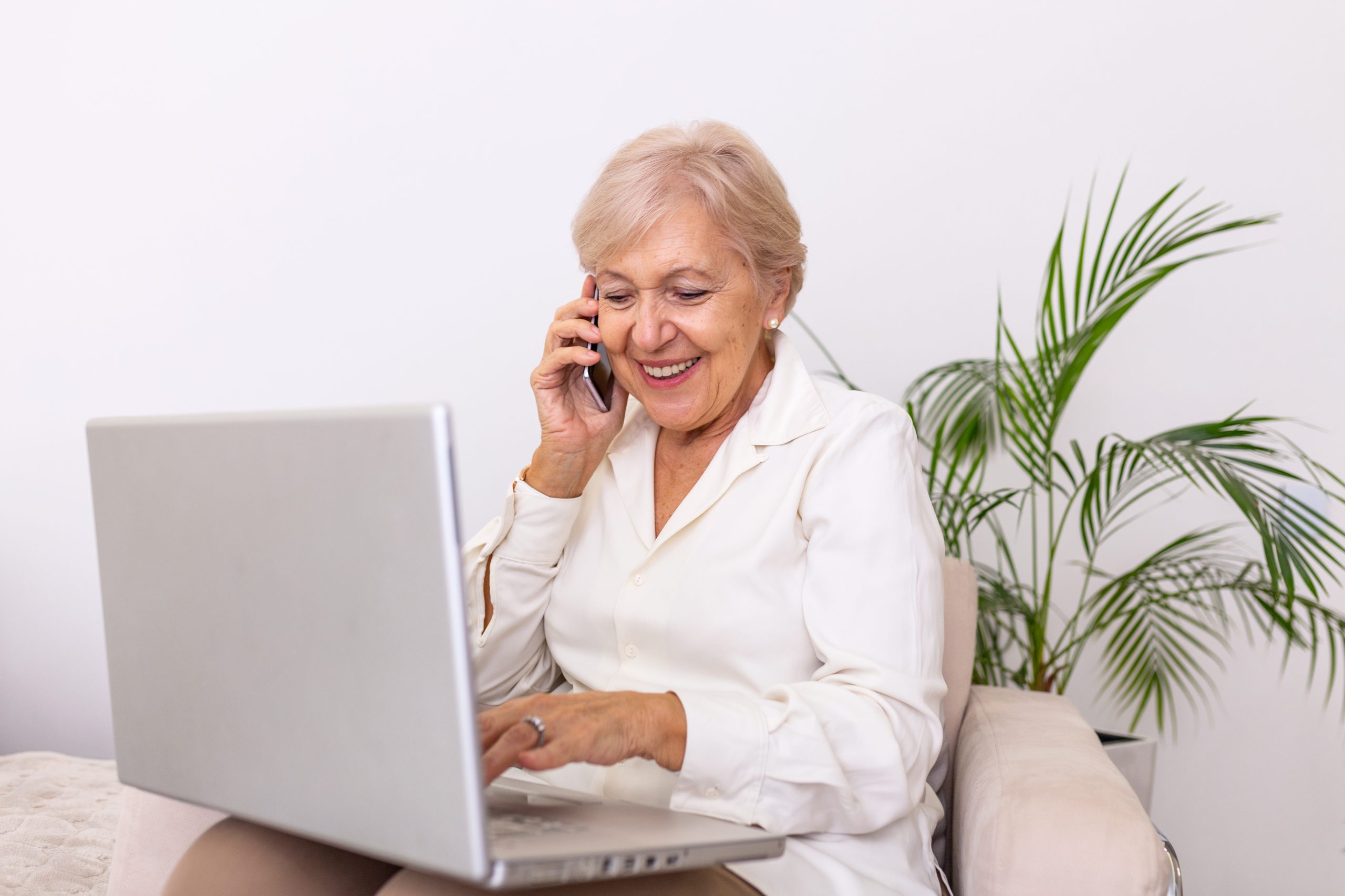 Elderly woman working on laptop computer, smiling, talking on the phone. Senior woman using laptop. Elderly woman sitting at home, using laptop computer and talking on her mobile phone, smiling. social media for the elderly