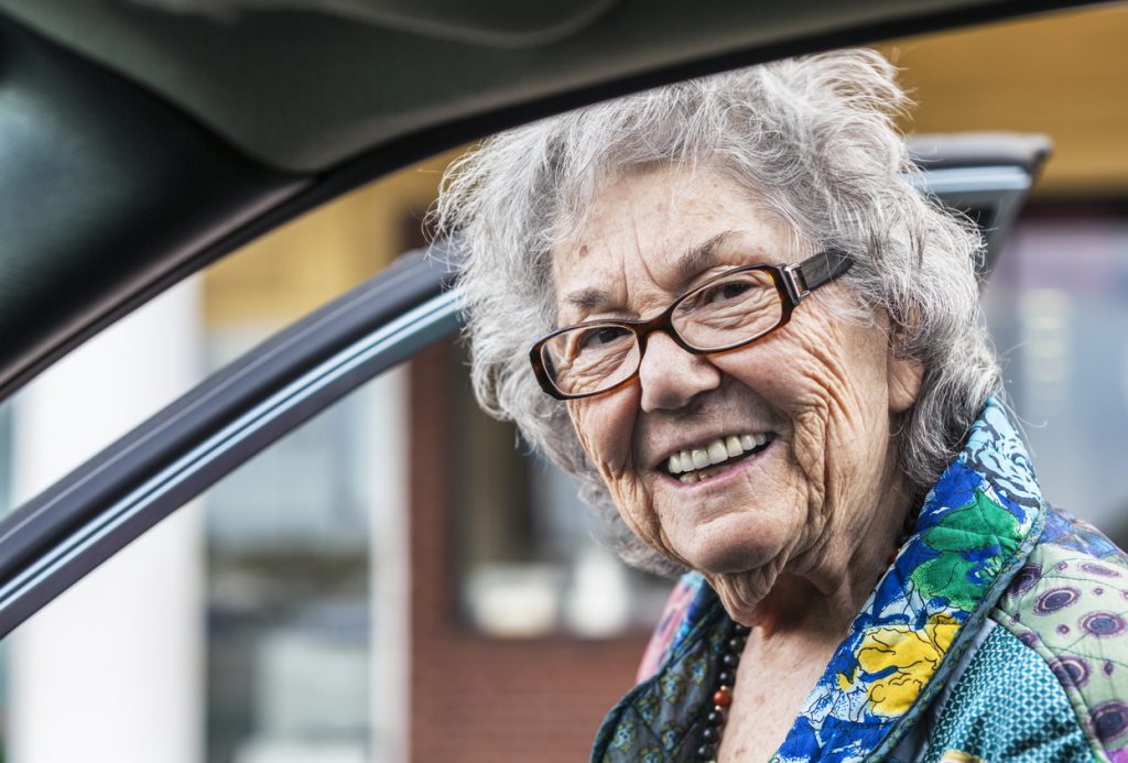 An elderly woman is smiling as she enters through the front passenger door of a car. Uber ride-sharing for seniors concept
