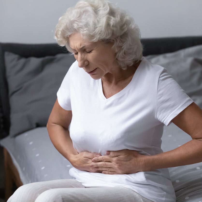 Elderly woman sits on her bed holding her stomach because of pain from a gastrointestinal disorder.