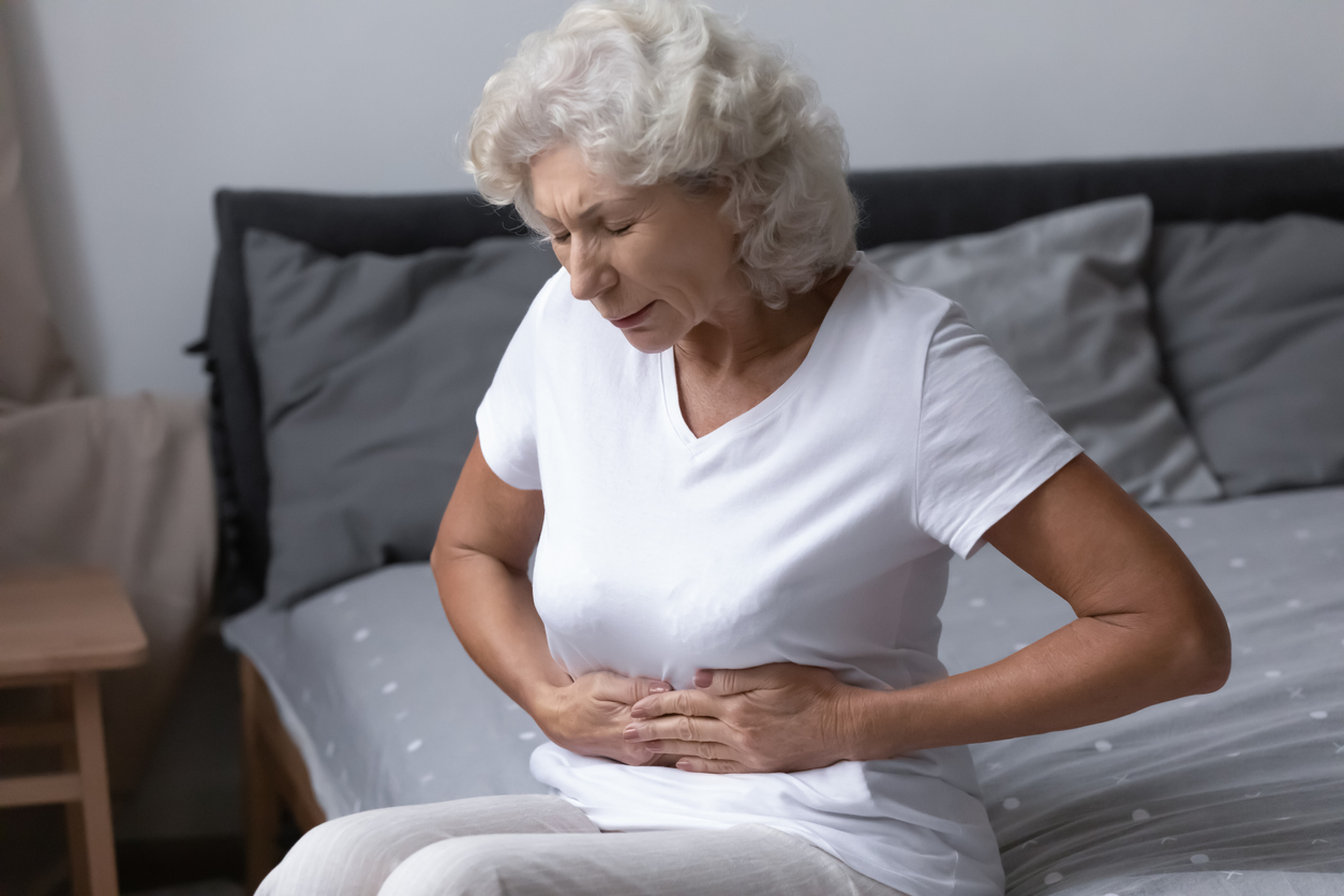 Elderly woman sits on her bed holding her stomach because of pain from a gastrointestinal disorder.
