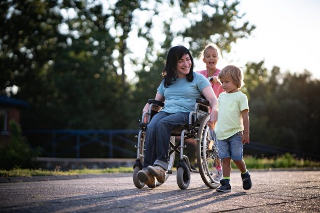 A mother with a spinal cord injury is spending time outside with her children.