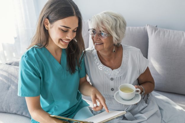 Happy elder woman sitting on white sofa and listening to nurse reading a book out loud home care agency and home health aides help aging in place.