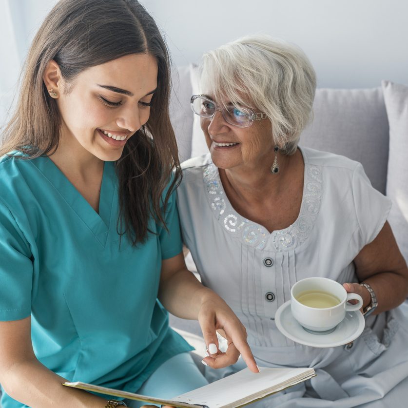 Happy elder woman sitting on white sofa and listening to nurse reading a book out loud home care agency and home health aides help aging in place.